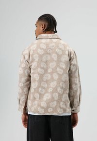 Beige jas met patroon en zachte textuur, met een paisley ontwerp, een kraag en manchetten met knopen. Gedragen over een wit overhemd en zwarte broek.