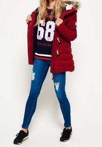 Czerwona pikowana parka z kapturem wykończonym sztucznym futrem, granatowa bluza z napisami w kolorze białym, podarte niebieskie dżinsy i czarne buty sportowe.