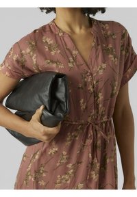 Robe rose poudré à motif floral, avec des manches courtes et un décolleté en V. Un portefeuille en cuir noir, avec des plis doux, est tenu dans une main.