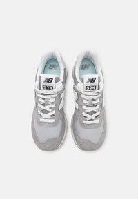 Pár šedobílých tenisek New Balance 574 s detaily z prodyšné síťoviny a semiše, bílými tkaničkami a světle modrými stélkami, zobrazených z pohledu shora.