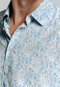 Korte mouwen shirt met een lichtblauw en crème bladeren patroon. Gemaakt van stof met een gladde textuur, voorzien van lichtgekleurde knopen.