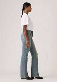 Γυναίκα με πλεξούδες φοράει λευκό μπλουζάκι, σκισμένα μπλε τζιν, μαύρες μπότες και ασημένια βραχιόλια, στεκόμενη σε προφίλ με ουδέτερο φόντο.