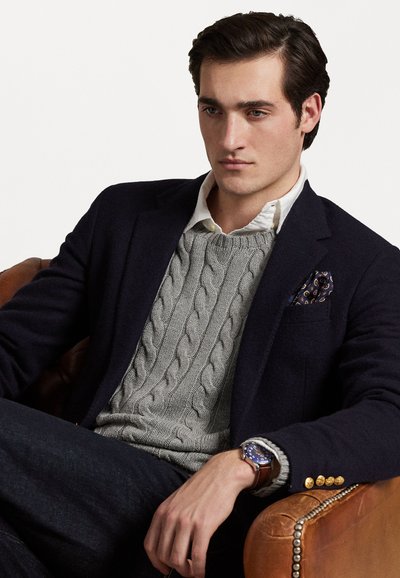 Blazer navy con botones dorados sobre un suéter de punto en gris, combinado con una camisa blanca. Finalizando con un reloj y un pañuelo de bolsillo de estampado.