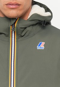Zelenoolivová nepremokavá bunda s kapucňou vystlanou sherpa, bielym zipsom a logom K-Way. Obsahuje viacfarebné pruhované detaily na zips.