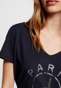 Camiseta de algodón azul marino de mangas cortas y escote en V. Presenta una impresión gráfica plateada de "PARÍS" y la Torre Eiffel.