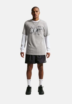 Hombre de pie vistiendo camisetas Nike superpuestas en gris y blanco, pantalones cortos Nike negros, calcetines blancos y zapatos deportivos negros sobre fondo blanco.