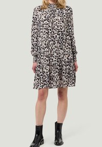 Femme portant une robe en imprimé léopard à manches longues avec des poignets smockés et des bottines noires, se tenant devant un fond clair uni.