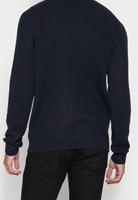 Man som bär en mörk marinblå ribbad tröja och svarta byxor, sedd bakifrån med avslappnade armar längs sidorna.