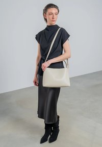 Weißes Leder-Umhängetasche mit glatter Textur, ausgestattet mit einem dezenten Logo und einem verstellbaren Tragegurt. Das Model trägt ein schwarzes Oberteil und einen Rock.