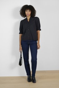 Zwarte blouse met korte mouwen, knoopsluiting en geplooide accenten, gecombineerd met donkerblauwe spijkerbroek en zwarte blokhaklaarzen. Houdt een kleine tas vast.