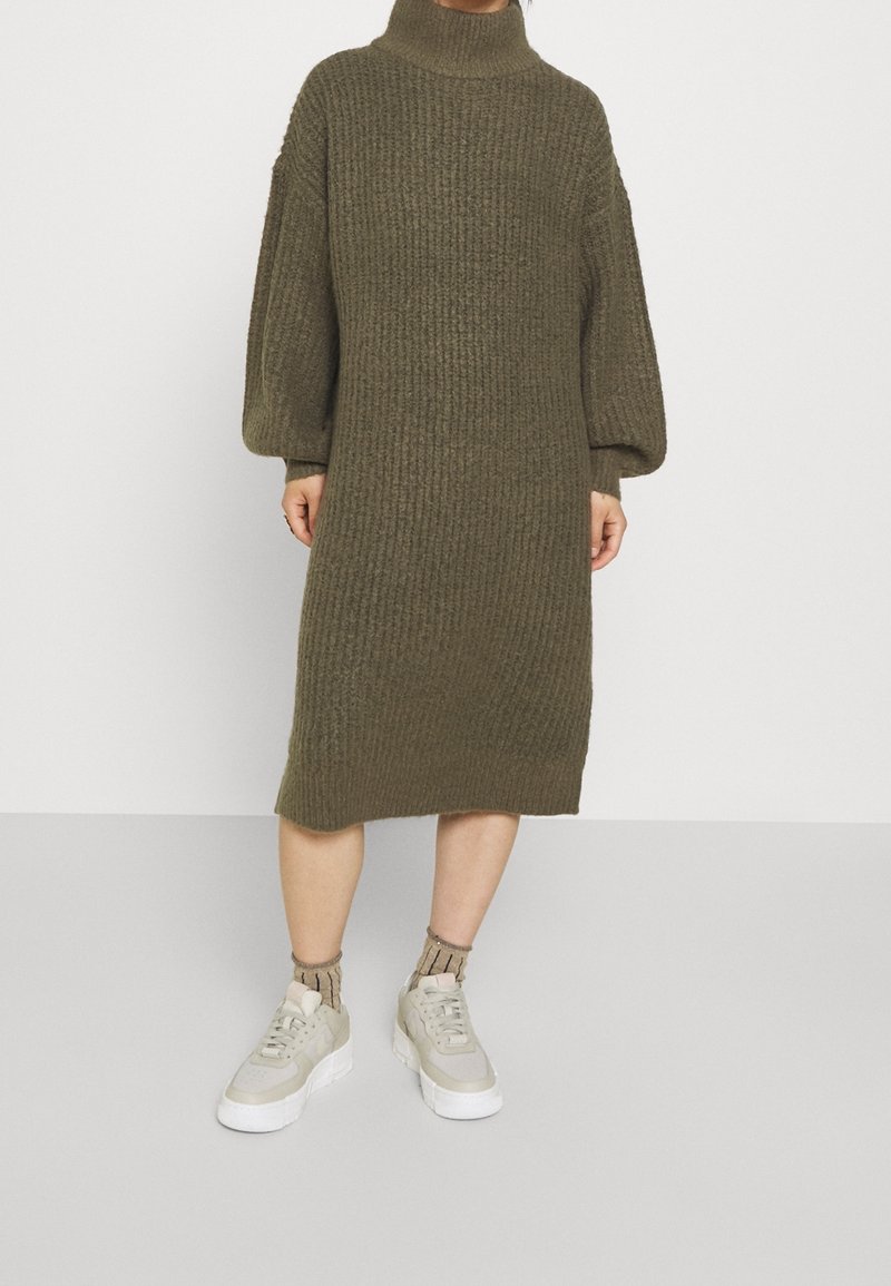 Personne portant une robe-pull en maille côtelée vert olive avec des manches bouffantes, associée à des baskets beiges et des chaussettes à motifs.