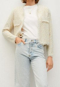 Frau trägt eine hellbeige strukturierte Cropped-Jacke, ein weißes Hemd, hellblaue high-waist Jeans und eine goldene Anhänger-Kette, die Hand in der Tasche.