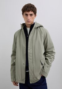 Nuori mies, jolla on lyhyet ruskeat hiukset, pitää yllään vaaleanvihreää hupullista takkia tumman vetoketjullisen neuleen ja sinisten farkkujen päällä, seisten yksinkertaista taustaa vasten.