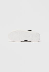 Zwarte sneaker liggend op zijn zijkant, met een witte zool waarin de merknaam "GUESS" in uiteenlopende groottes is gedrukt.