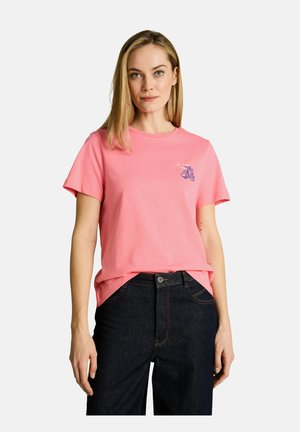 Vrouw draagt een roze T-shirt met een klein blauw scooterafbeelding en donkerblauwe spijkerbroek, staand tegen een effen witte achtergrond.