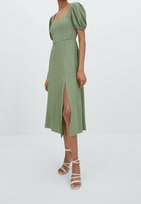 Robe verte claire, longueur genoux, avec un tissu texturé, décolleté carré et manches courtes bouffantes. Une fente sur le côté ajoute un détail ; associée à des talons à lanières.