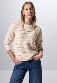 Cremfarbener Strickpullover mit strukturiertem Muster und Rundhalsausschnitt, kombiniert mit blauen Jeans. Weicher Stoff mit gerippten Bündchen.