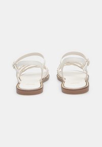 Valkoiset hihnasaappaat, joissa on litteät ruskeat pohjat, varustettu säädettävillä nilkkavöillä ja minimalistisella muotoilulla. Sileä pinta, jossa on hillittyä yksityiskohtaisuutta.