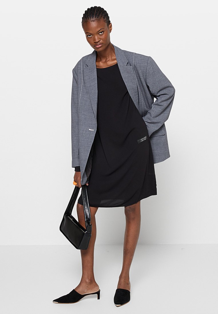 Abito nero senza maniche sotto un blazer grigio oversize, abbinato a scarpe slip-on nere e a una piccola borsa nera. Tessuto morbido e design strutturato.
