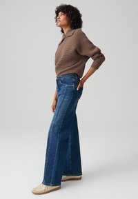 Pullover marrone a coste con maniche ampie, abbinato a jeans a vita alta blu svasati. Presenta una vestibilità dritta e leggere scoloriture all'orlo.