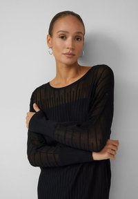 Jeune femme avec des cheveux tirés en arrière, portant des boucles d'oreilles en argent et un haut noir transparent à manches longues avec des côtes, les bras croisés sur la poitrine.