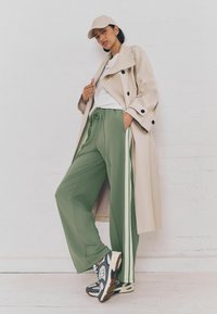 Beige trench coat mustade nuppudega, rohelised dressipüksid valgete külgtriipudega ja hallid tossud. Mudel kannab beeži mütsi ja valget särki.
