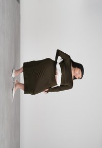 Femme en robe longue à manches longues vert olive foncé avec une ceinture blanche et des chaussures à talons blancs posant devant un mur gris uni.