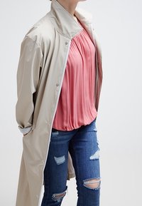 Długi beżowy płaszcz na różowej bluzce i wyblakłych niebieskich dżinsach. Płaszcz ma wysoki kołnierz i zatrzaski.