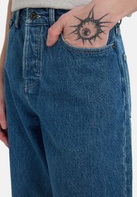 Jeans de mezclilla azul con un ajuste relajado, que presentan una cintura alta, un bolsillo frontal y una sutil textura desgastada. Cierre de botón negro visible.