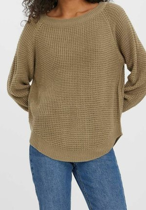 Kvinde iført en løs, lysebrun strikket sweater med tekstureret mønster og buet kant, kombineret med mellemblå jeans.