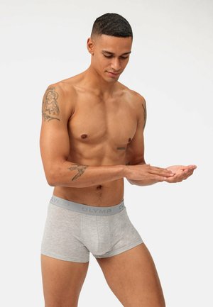 Graue Boxershorts aus weichem Material, mit einem grauen Bund, auf dem das Logo "OLYMP" zu sehen ist. Das Modell hat einen muskulösen Körperbau und gestikuliert mit seinen Händen.