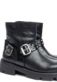 Botines de cuero negro hasta el tobillo con una correa ancha que presenta herrajes y tachuelas de plata, superficie texturizada y una suela gruesa negra.