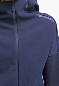 Marinblå zip-up jacka med texturerad tyg, huva och kontrasterande vertikala ränder längs axlarna.