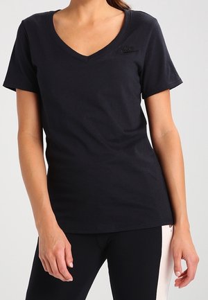 Femme portant un T-shirt noir Nike ajusté à col en V et des leggings noirs avec des panneaux latéraux blancs, se tenant devant un fond uni.