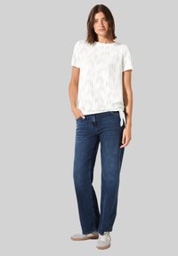 T-shirt bianca a maniche corte con design texturizzato, abbinata a jeans dritti blu scuro e sneakers grigie chiare. Il modello è in piedi con le mani nelle tasche.