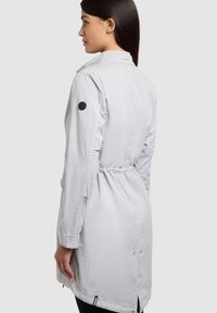 Femme aux longs cheveux foncés tournée de profil, portant un trench-coat gris clair à manches longues avec une taille cintrée et un patch rond noir sur le bras.