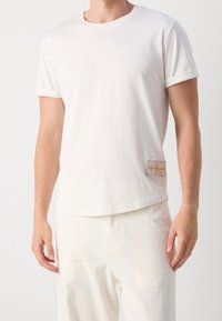 Vit bomulls-T-shirt med kort uppvikta ärmar, klassisk rund halsringning och en rektangulär etikett på vänster sida med varumärkesnamn och logotyp.