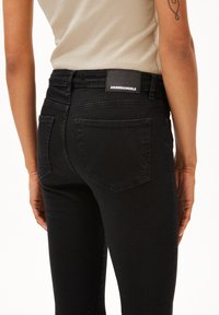 Jeans en denim noir à taille haute, poches arrière avec des coutures courbées, et une étiquette en cuir noir avec marque sur la ceinture. Texture douce.