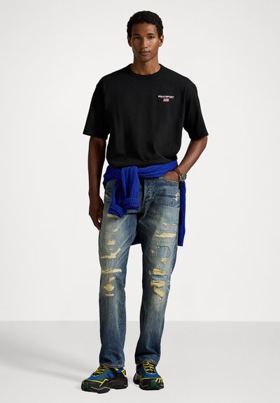 Camiseta de algodón negra con logo, suéter de punto azul atado a la cintura, jeans de mezclilla desgastados y zapatillas deportivas azul y negra.
