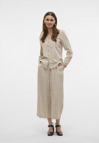 Donna in piedi con le mani nelle tasche, che indossa una camicia beige a righe con bottoni e pantaloni larghi coordinati, accompagnati da scarpe con tacco nere.