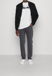 Chaqueta bomber negra con cremallera y bolsillos, camiseta blanca con texto negro, jeans grises y zapatillas blancas. Corte estándar con un estilo casual.
