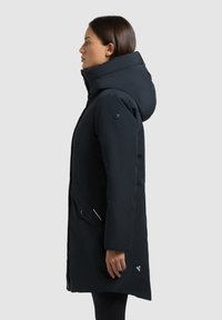 Mørkeblå parka med stor hætte, frontknapper og sidelommer. Har en glat tekstur og et logo-accent nær kanten.