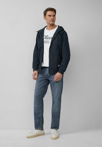 Granatowa, lekka kurtka z kapturem, z przednimi kieszeniami i elastycznymi mankietami, noszona na białej koszulce z grafiką, w zestawieniu z jasnoniebieskimi dżinsami i trampkami.