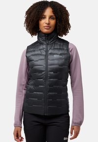 Schwarze, gepolsterte Weste mit einem hohen Kragen und Reißverschluss, die ein strukturiertes lila Langarmshirt darunter und schwarze Hosen trägt.