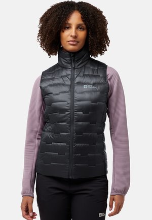 Zwarte gewatteerde vest met een hoge kraag en rits aan de voorkant, met daaronder een getextureerd paarse blouse met lange mouwen en zwarte broek.
