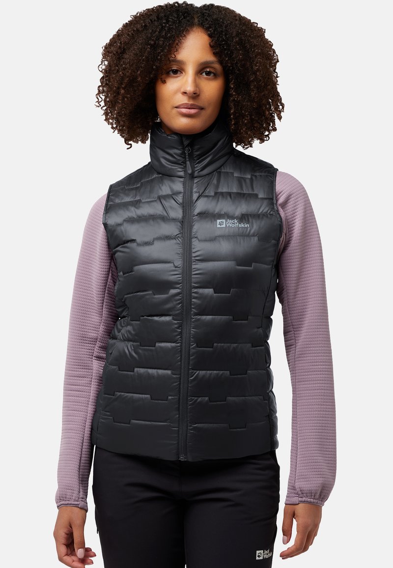 Schwarze, gepolsterte Weste mit einem hohen Kragen und Reißverschluss, die ein strukturiertes lila Langarmshirt darunter und schwarze Hosen trägt.