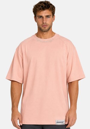 Jeune homme aux cheveux bouclés portant un t-shirt ample rose clair à manches courtes avec une petite étiquette près de l'ourlet et un texte subtil sur le col.
