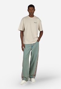 Beżowa, oversize'owa koszulka z haftowanym napisem, w zestawie z jasnoniebieskimi spodniami o szerokich nogawkach z czerwonymi i białymi paskami po bokach oraz białymi trampkami.