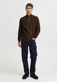 Brown zip-neck sweater with a textured finish, paired with dark blue denim jeans and black shoes, displayed on a neutral background.