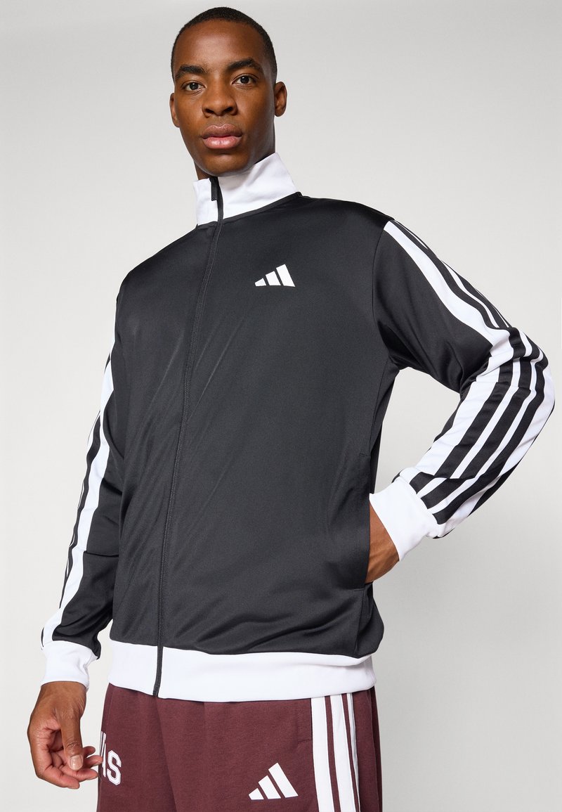 Chaqueta negra con cremallera y rayas blancas en las mangas, con un logo blanco en el lado izquierdo del pecho y puños y dobladillo blancos.